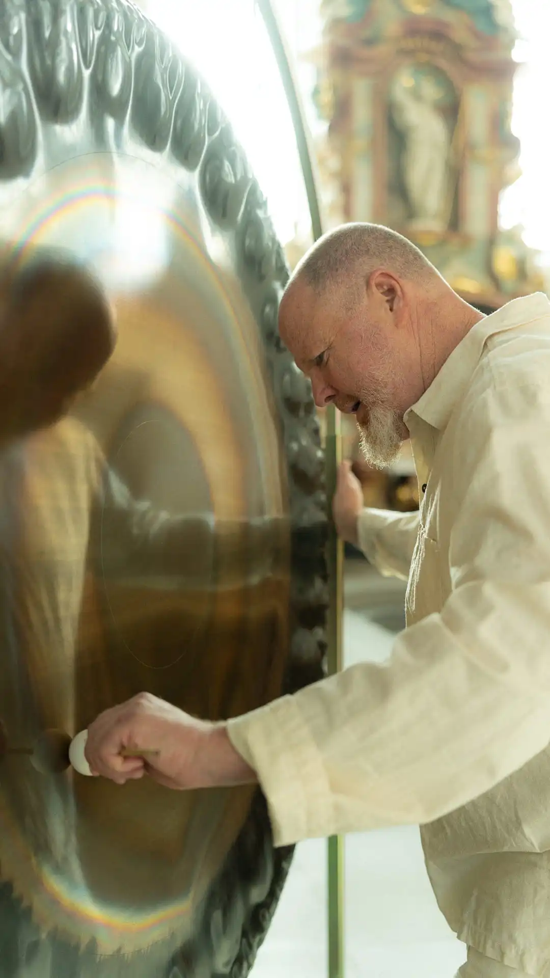 Man in light clothing playing a large gong with a mallet in an indoor setting.
