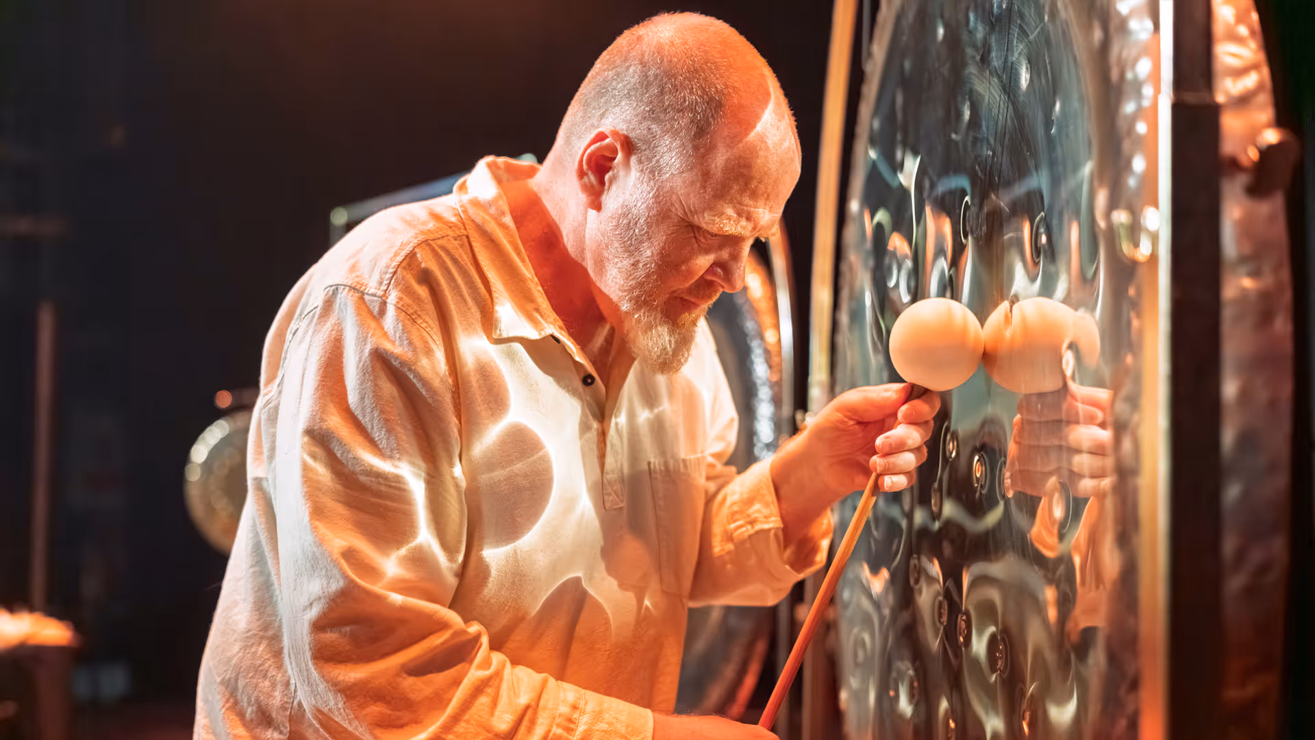 Man in white shirt playing a large hammered gong with mallets in a dimly lit room.