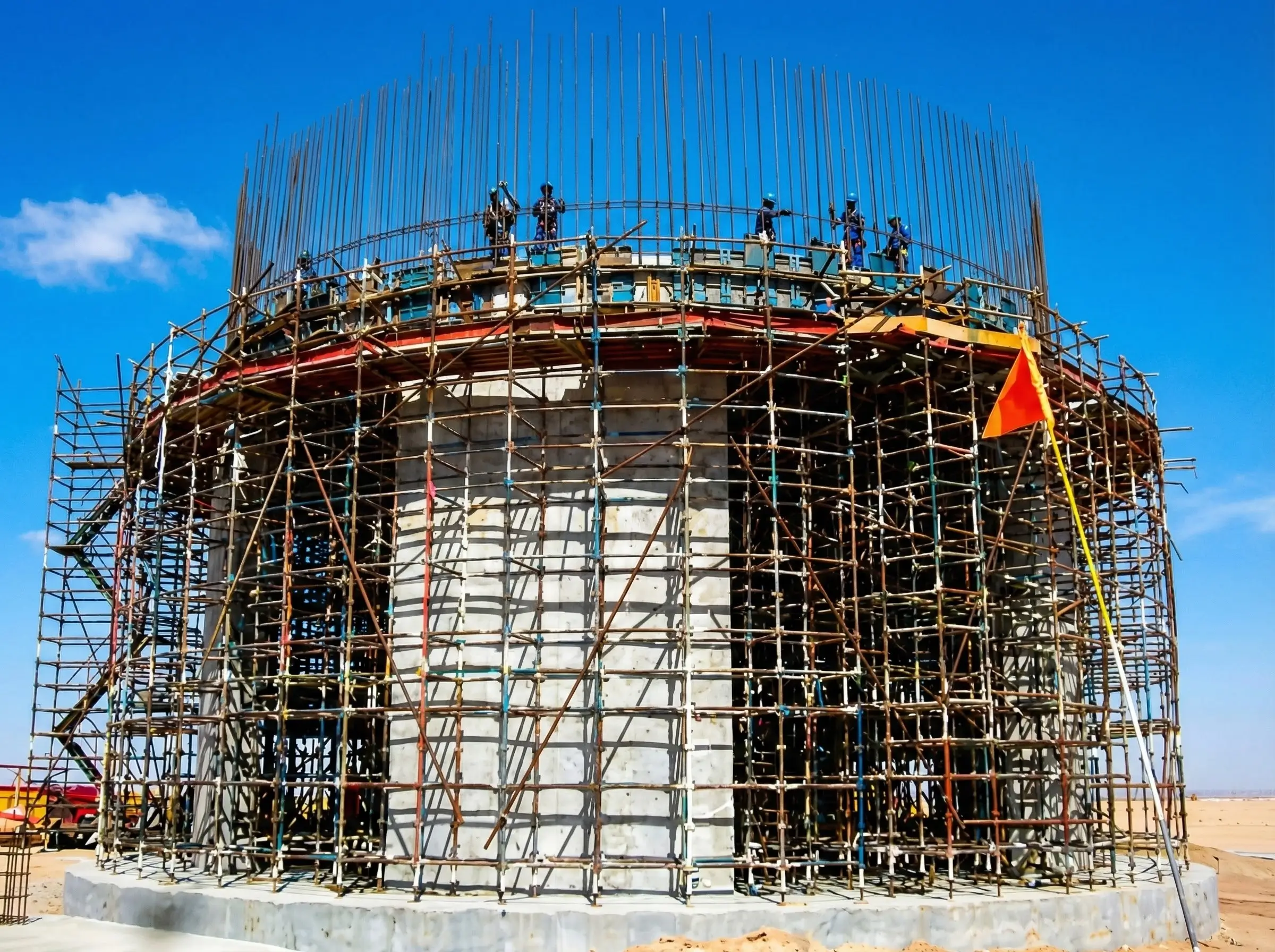 Circular concrete structure under construction with scaffolding and workers on top against a clear blue sky.