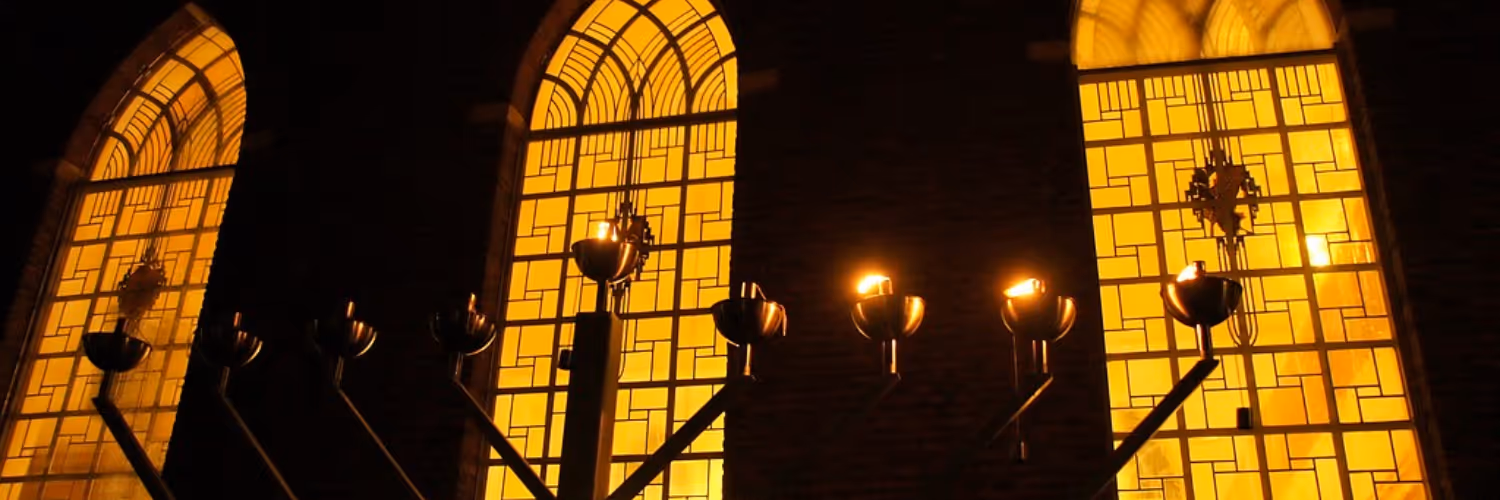 Gloeiende traditionele kandelaar met brandende vlammen voor geel verlichte gebrandschilderde ramen in een donkere ruimte.