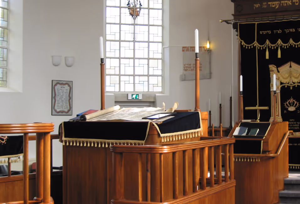 Interieur van een synagoge met een houten lezenaar bedekt met een zwarte doek en boekrollen, naast andere houten meubelen en kaarsen.