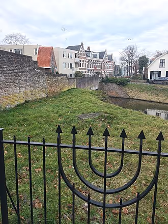 IJzeren hek met menora voor een groene begraafplaats met historische huizen en muren op de achtergrond.