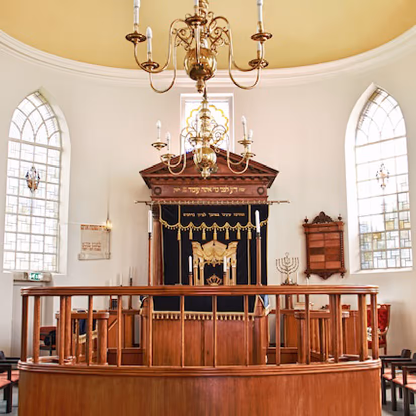 Interieur van een synagoge met houten hekwerk, twee hoge ramen en een geborduurd altaar met kandelaar en kroonluchters.