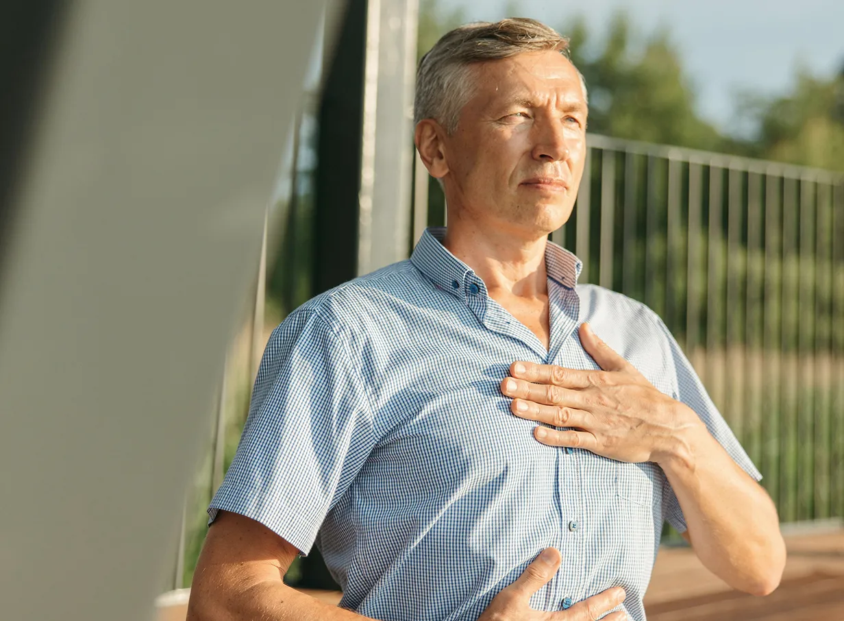 A man sitting on a wooden deck outdoors with his eyes closed and hands on his chest and stomach, practicing deep breathing.