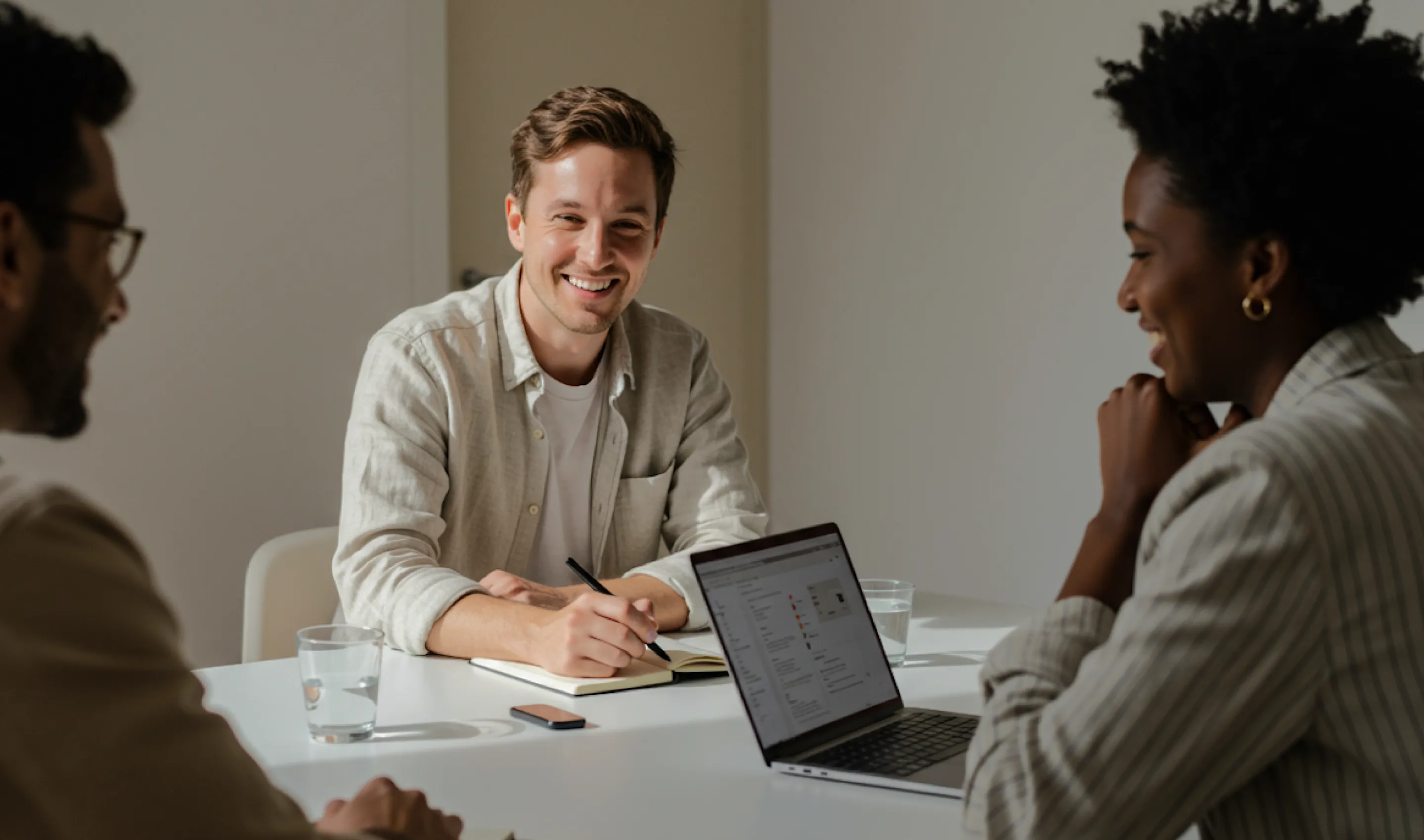 A group of professionals in an office setting, with a man smiling and taking notes during a meeting.