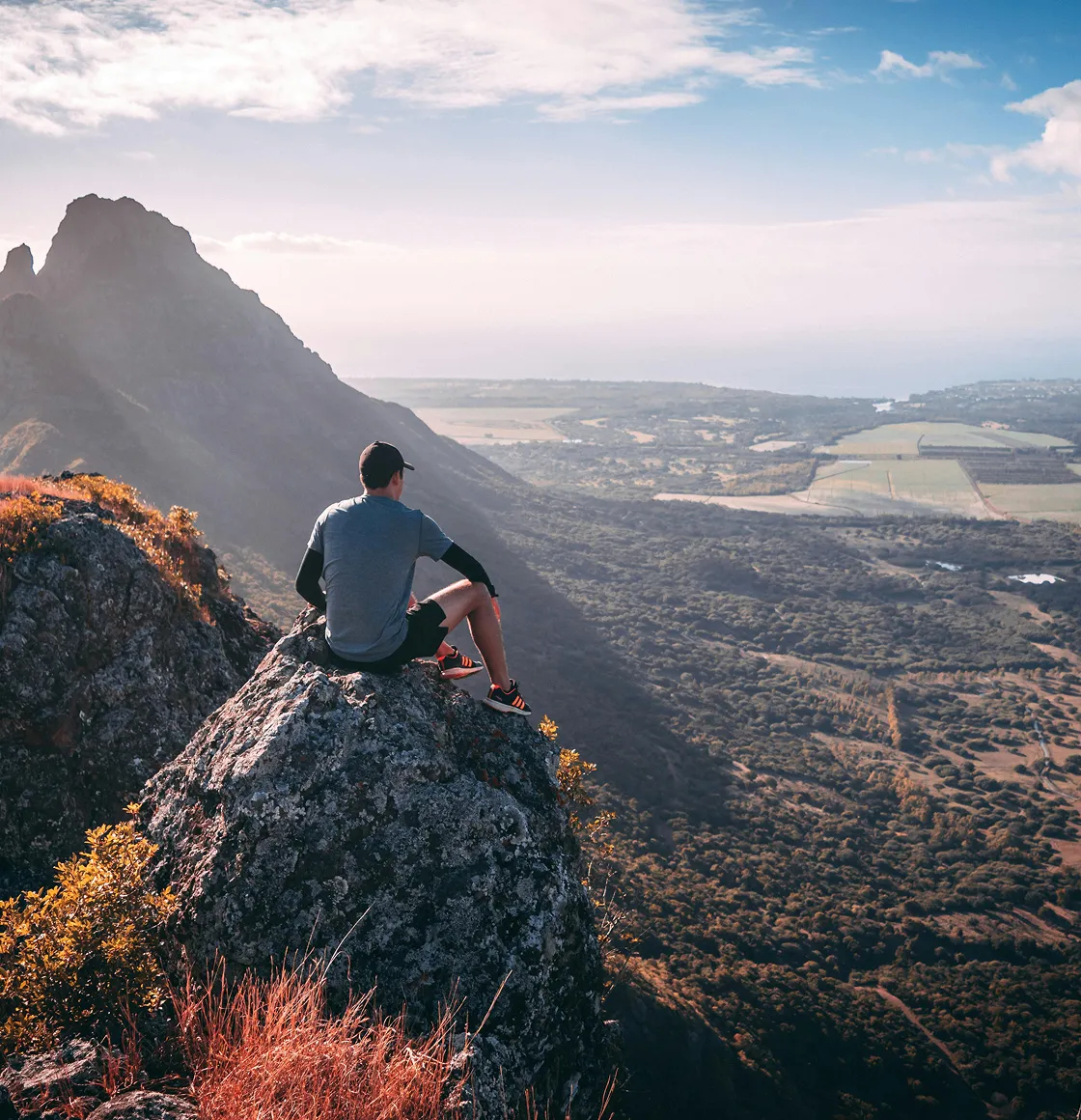 A man sits on a rocky peak overlooking a vast green valley and the ocean.
