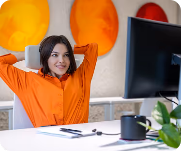 Smiling woman in orange shirt leaning back with hands behind her head at a white desk with a computer monitor.