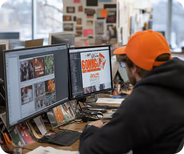 Person in orange cap working on graphic design with dual monitors displaying images and text.
