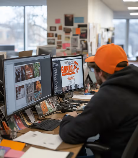 Person wearing an orange cap working on a dual-monitor computer setup displaying graphic design and photography content in an office.
