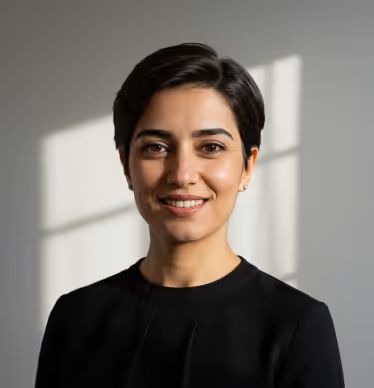 Smiling woman with short dark hair wearing a black top, standing in front of a window casting a shadow on a gray wall.