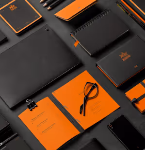 Flat lay of black and orange branded stationery including notebooks, pens, glasses, smartphone, and a leather folder.