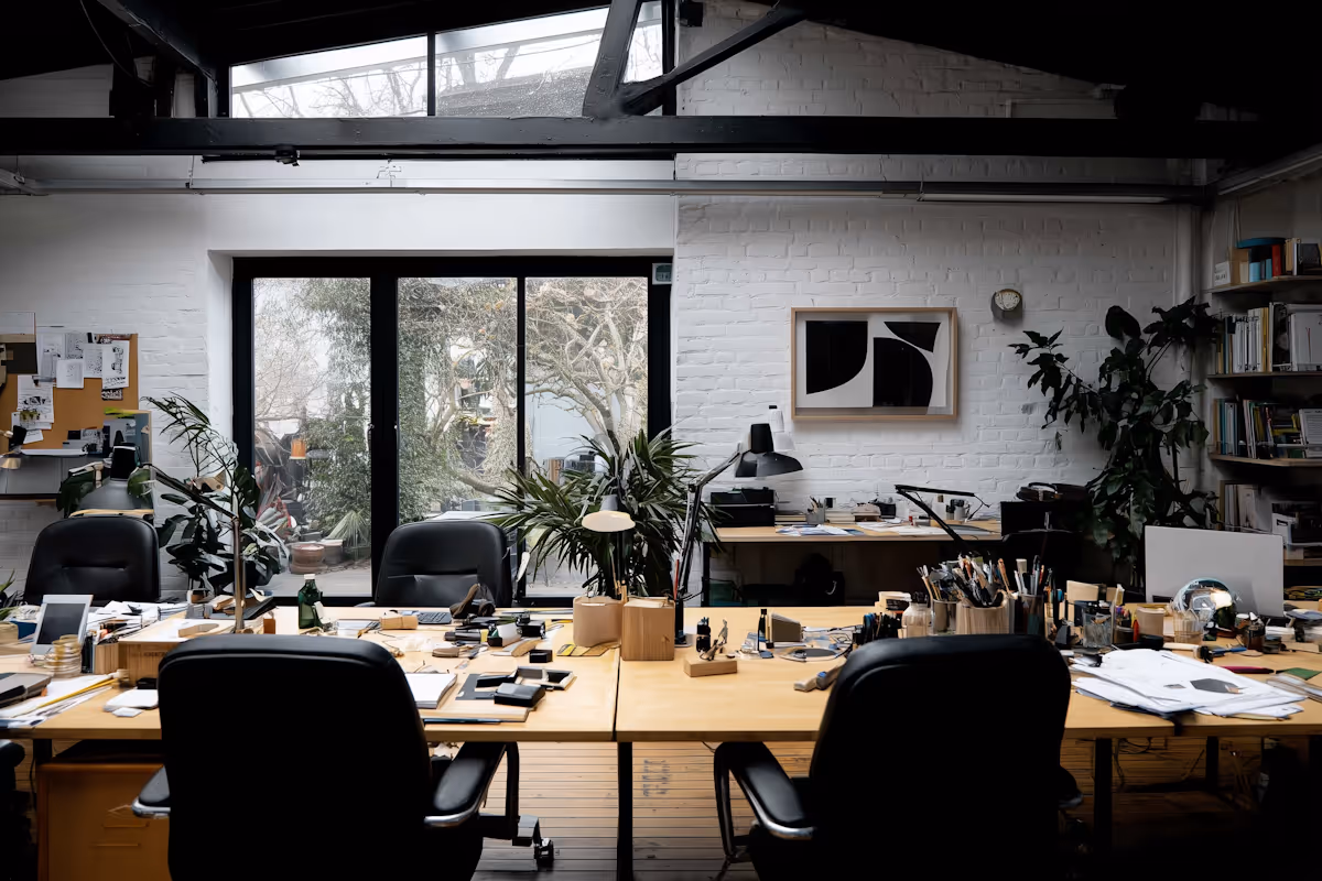 Modern creative office with wooden desks, black chairs, plants, desk lamps, and large window overlooking trees.