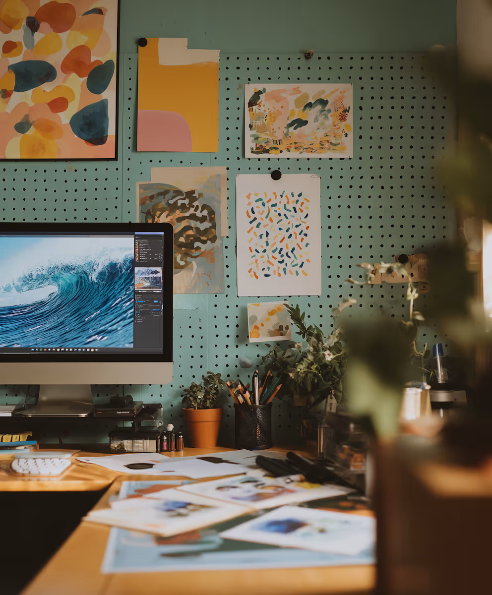 Creative workspace with a desktop computer showing a wave photo editing, colorful abstract artwork on a pegboard wall, and various stationery on the wooden desk.
