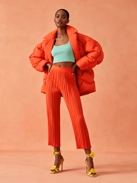 Woman wearing a bright orange puffer jacket, matching pleated orange pants, a light green cropped top, and yellow high-heeled sandals with flower details, standing against a peach background.