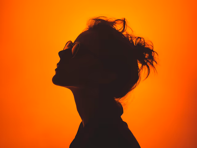 Silhouette of a woman wearing sunglasses against a bright orange background, her hair tied up in a messy bun.