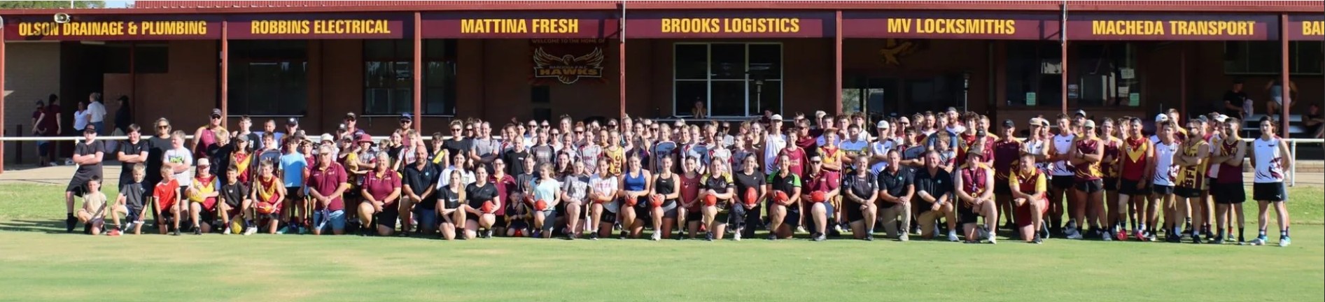 Large group of people posing on a grass field in front of a building with sponsor signs overhead.