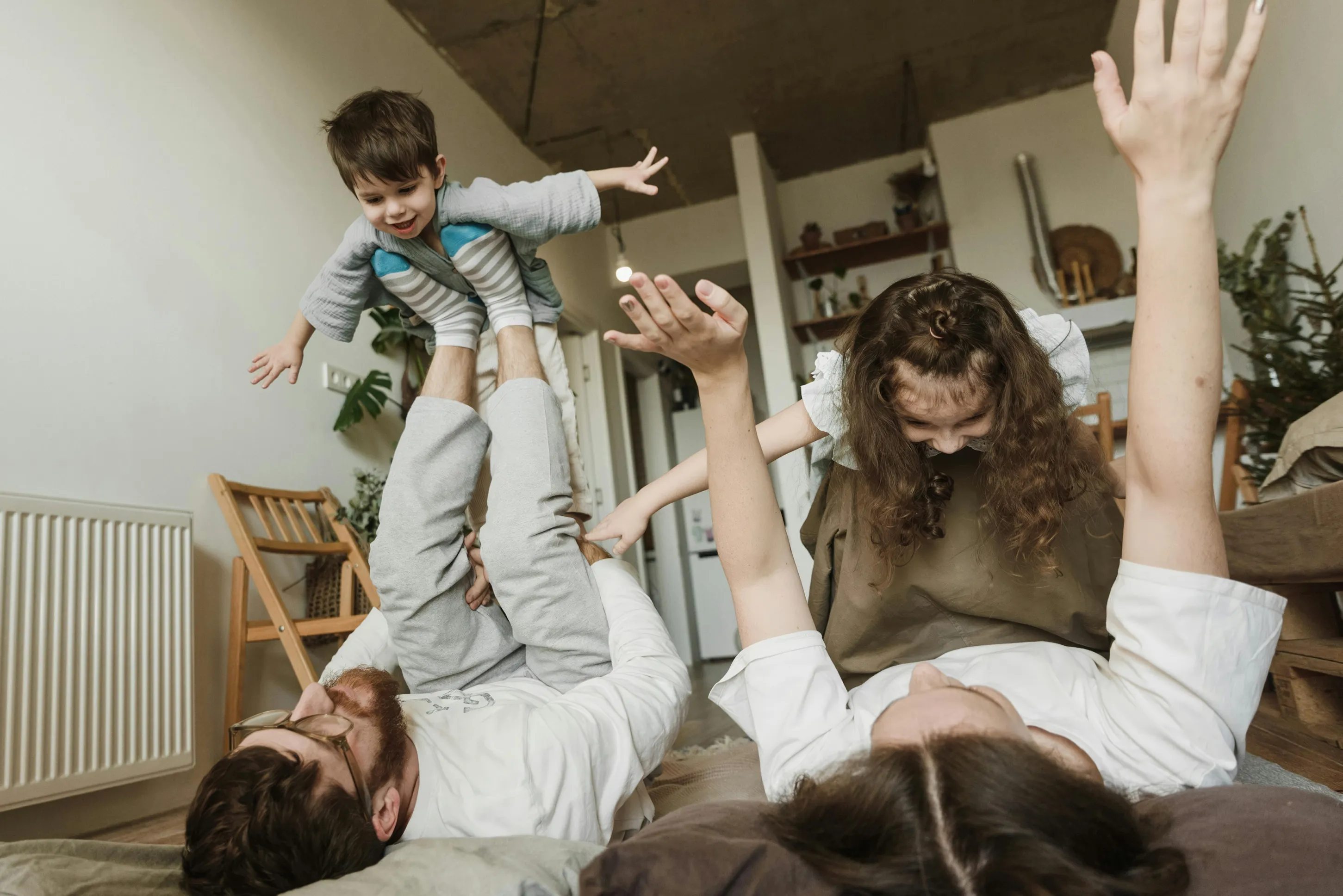 Parents lying on the floor playfully lifting a smiling child and a girl in an indoor living room.