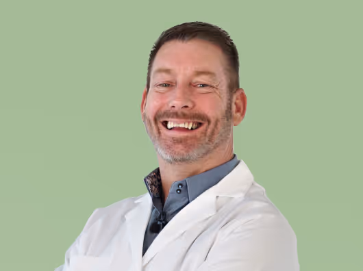 Smiling man with short hair and beard wearing a white lab coat and blue shirt against a green background.