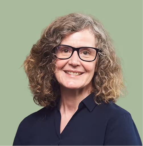 Smiling older woman with curly gray hair and black glasses wearing a dark blue shirt against a muted green background.
