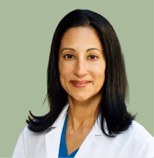Woman with dark hair wearing a white lab coat and blue top, smiling against a green background.