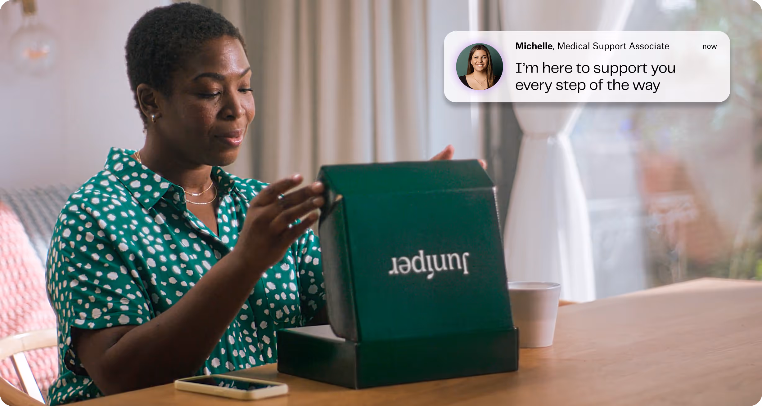 Woman in green patterned shirt opening a green Juniper box at a wooden table with a smartphone nearby.