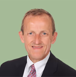 Professional headshot of a middle-aged man with short light brown hair, wearing a black suit, white shirt, and patterned tie with a green background.
