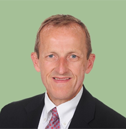 Professional headshot of a middle-aged man with short light brown hair, wearing a black suit, white shirt, and patterned tie with a green background.