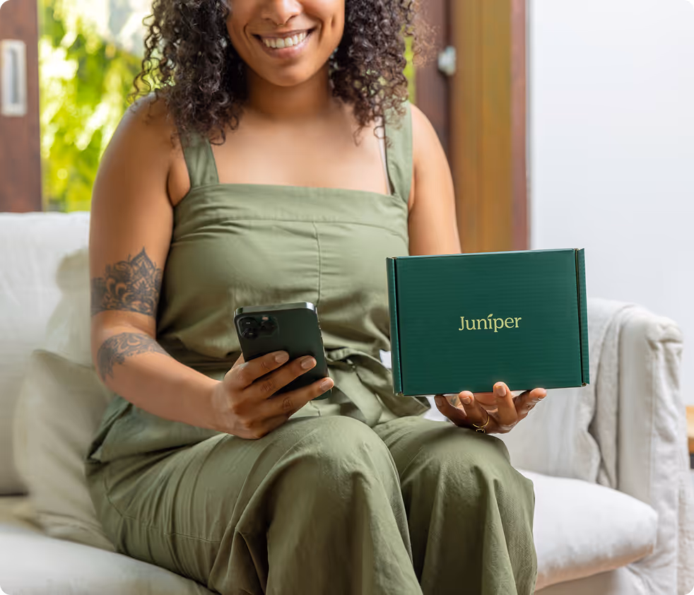 Smiling woman in green outfit sitting on a couch holding a smartphone and a green box labeled 'Juniper'.