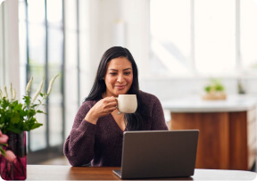 Woman sitting at a table using a laptop while holding a white coffee mug.