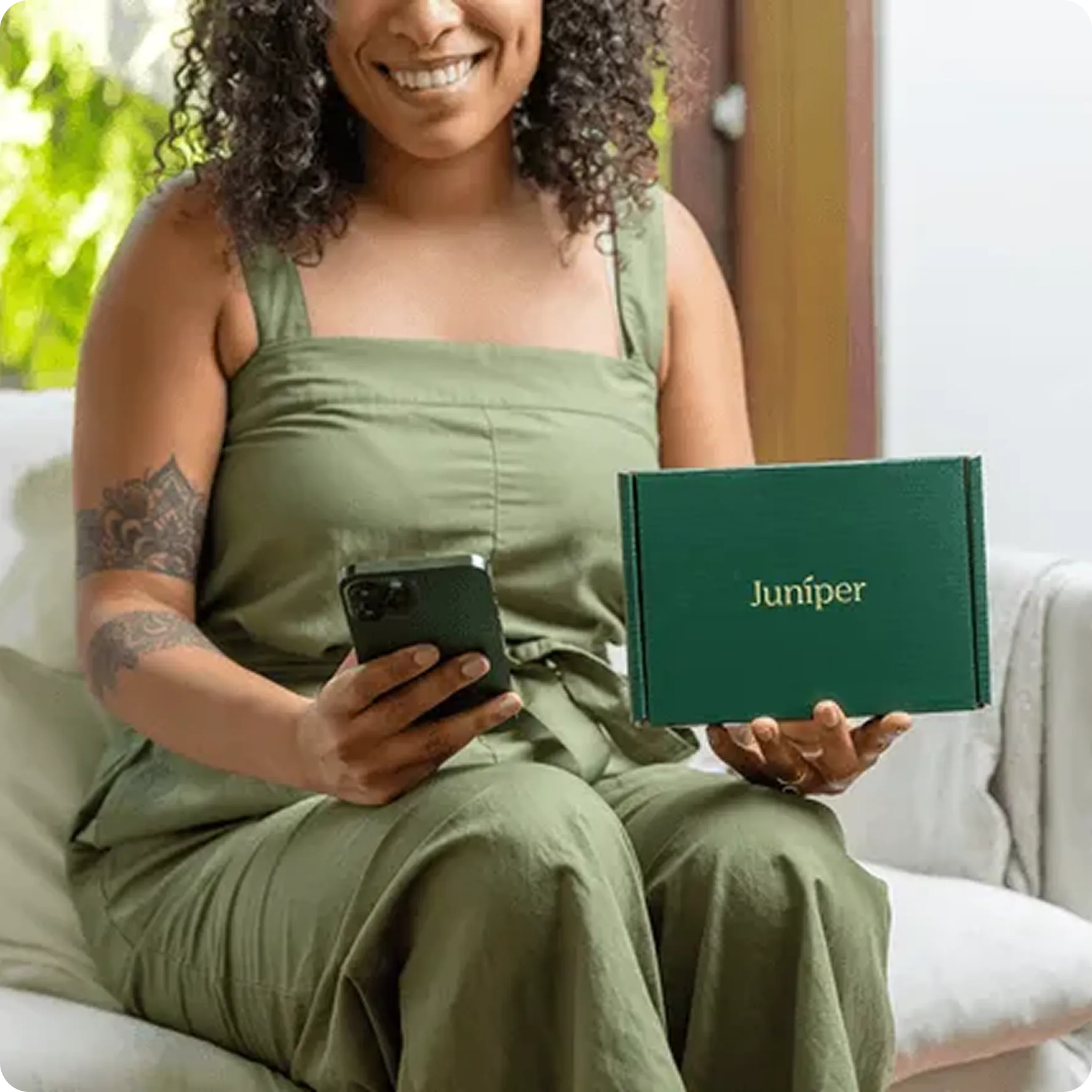 Smiling woman in green outfit holding a phone in one hand and a green box labeled Juniper in the other.
