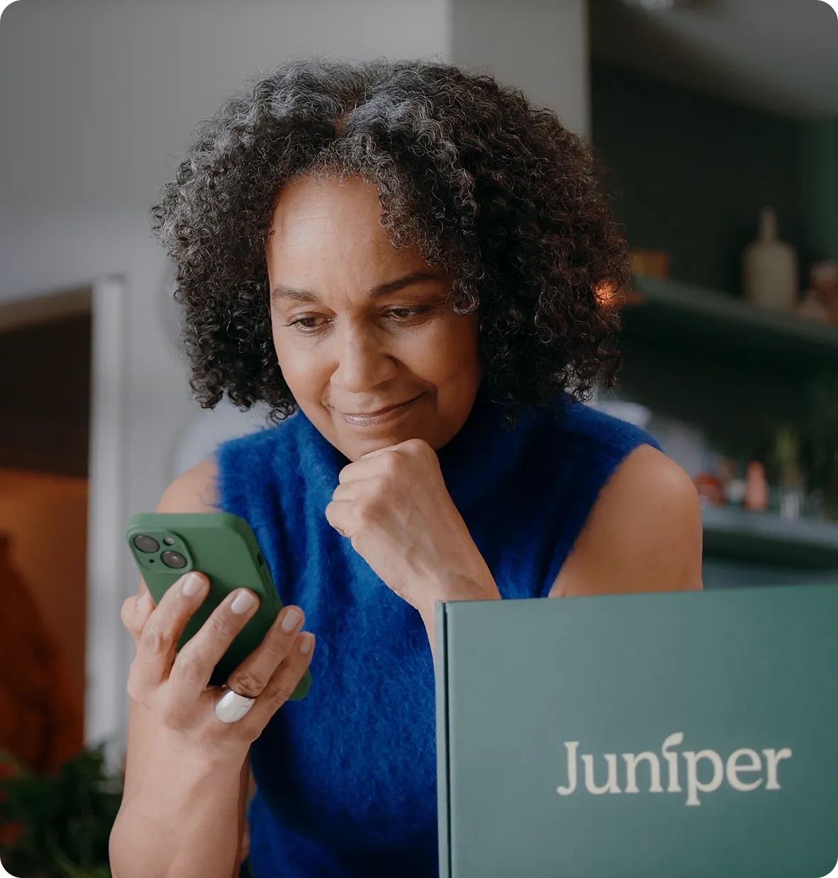Woman with curly hair wearing blue sleeveless top looking at smartphone and smiling softly.