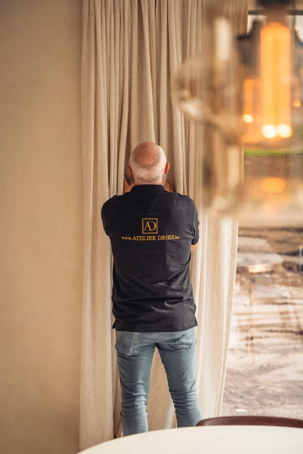 Man met grijs haar, gekleed in een zwart Atelier Dries-shirt, die voelt aan beige gordijnen in een kamer.