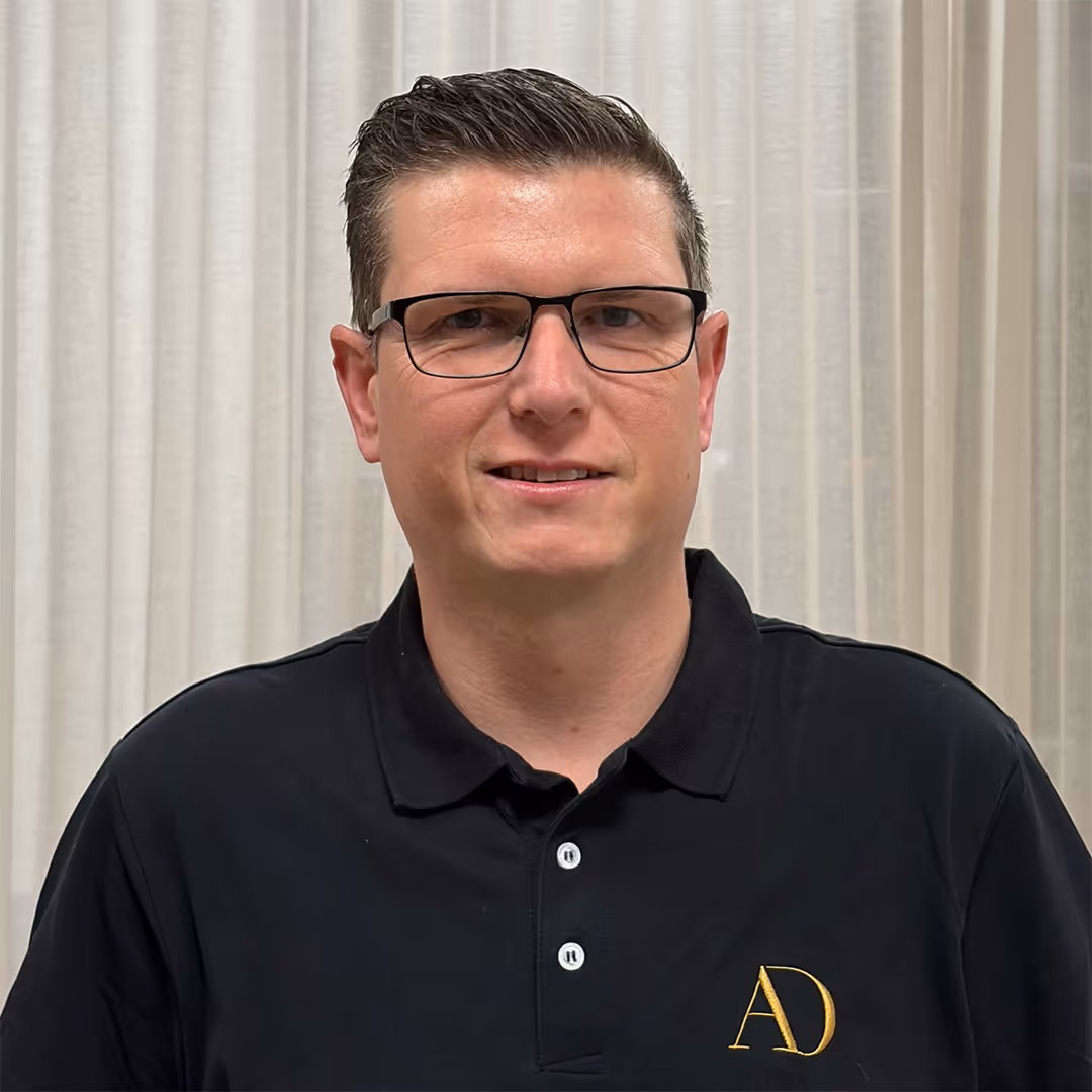 Glimlachende man met bril en bruin haar, gekleed in een zwart poloshirt met gele AD-initialen, staande voor vitrages.