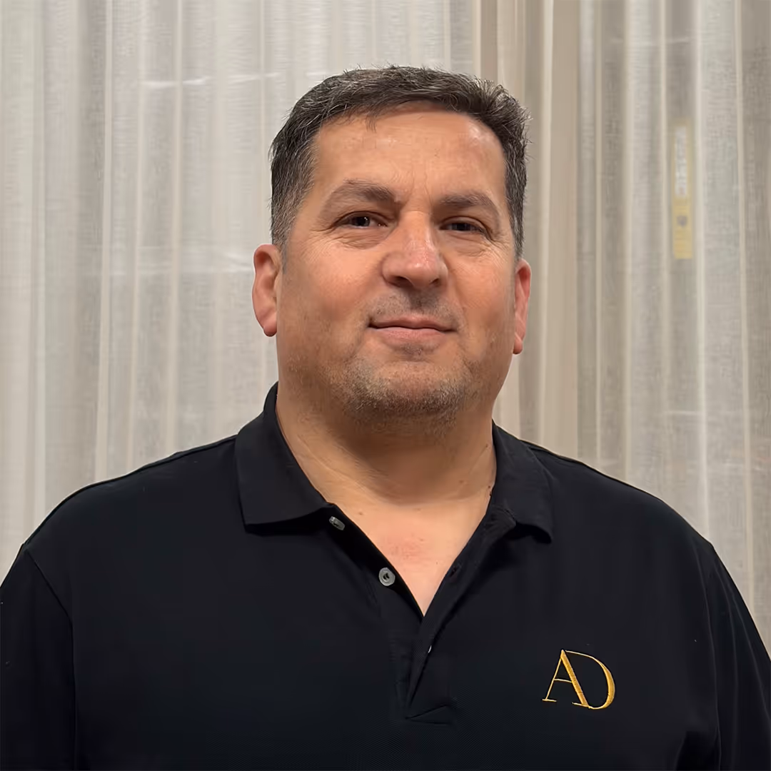 Glimlachende man van middelbare leeftijd met bruin haar, gekleed in een zwart poloshirt met gele AD-initialen, staande voor vitrages.