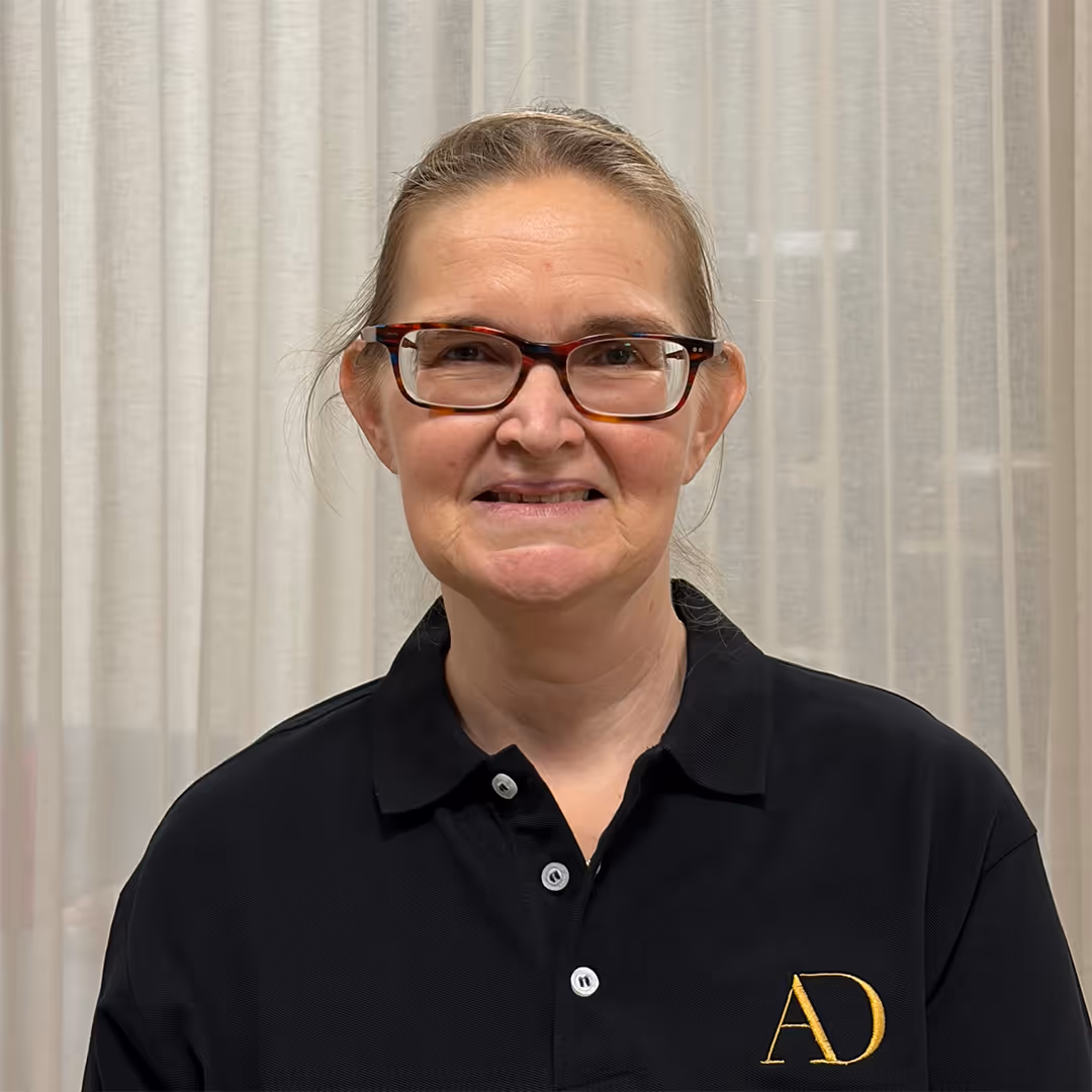 Glimlachende vrouw van middelbare leeftijd met bril, gekleed in een zwart poloshirt met gele AD-initialen, staande voor vitrages.