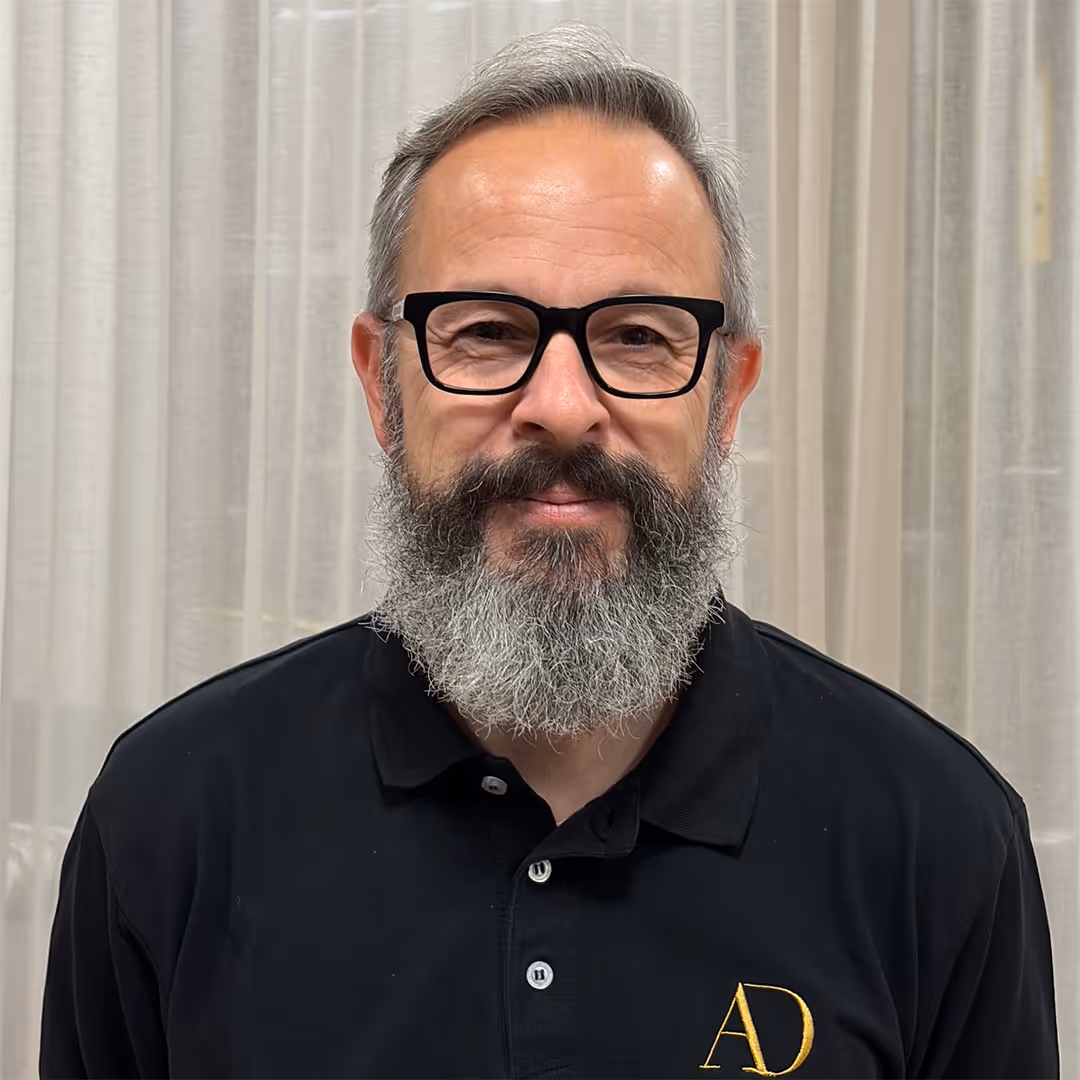Man van middelbare leeftijd met grijze baard, bril en zwart poloshirt met gouden AD-logo voor een beige gordijn.