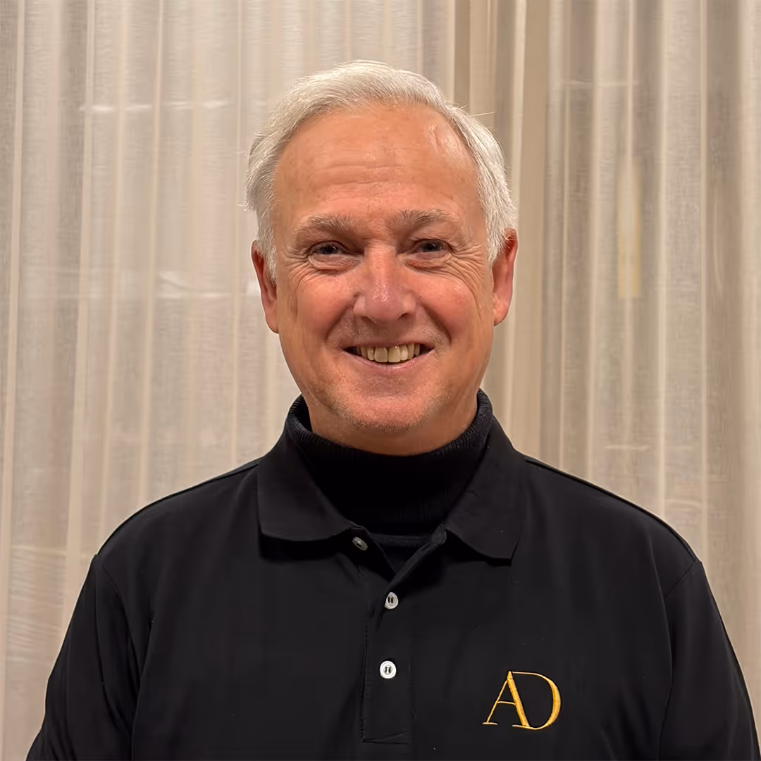 Glimlachende oudere man met wit haar, gekleed in een zwart poloshirt met een gouden AD-logo.