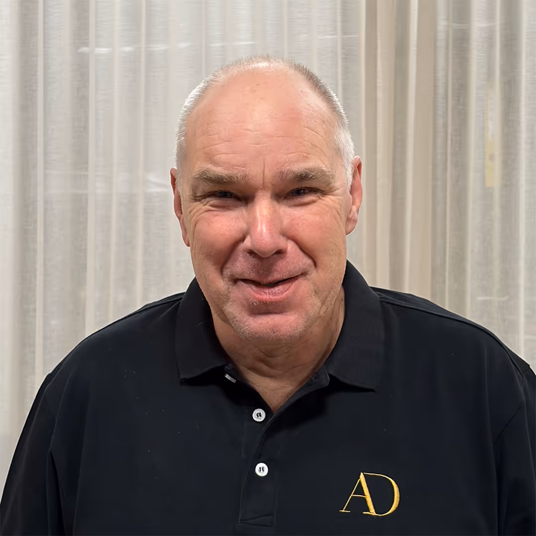 Glimlachende, kale man van middelbare leeftijd met bril, gekleed in een zwart poloshirt met gele AD-initialen, staande voor vitrages.