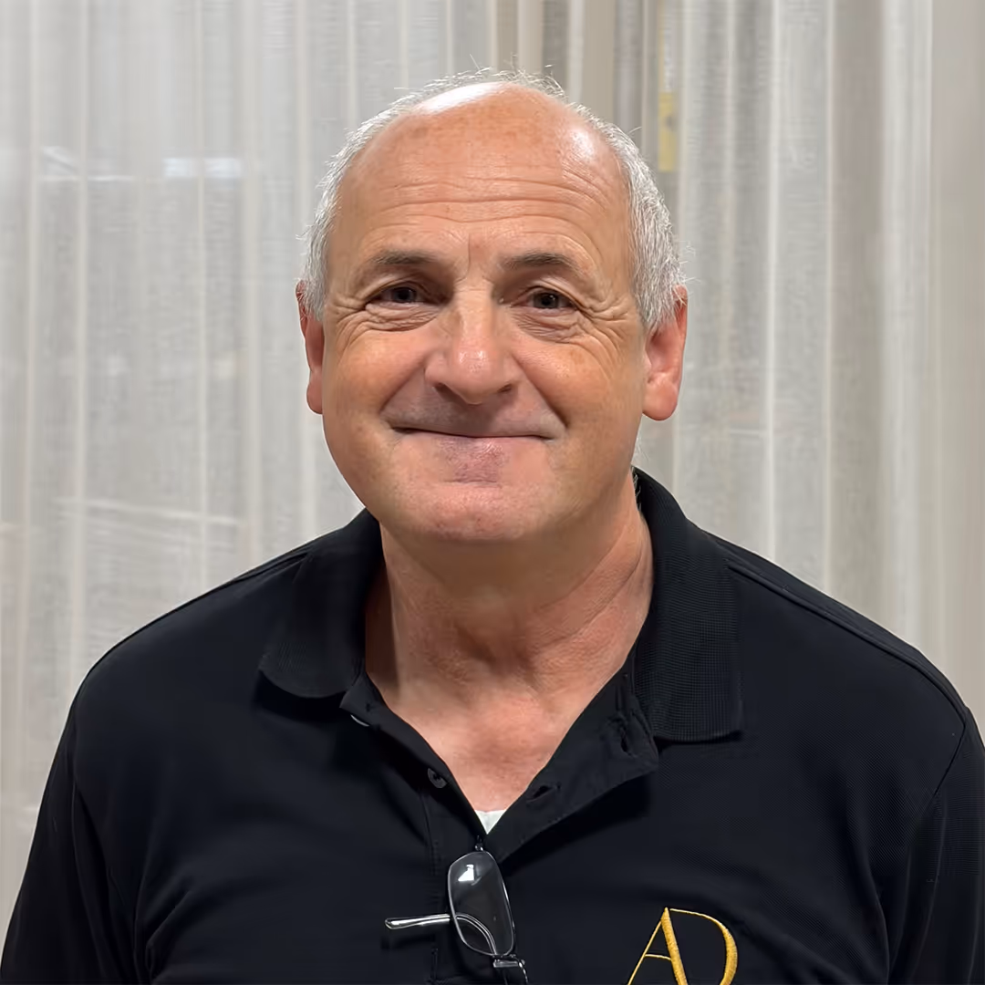 Glimlachende oudere man met kort grijs haar, gekleed in een zwart poloshirt met een gouden AD-logo en een bril die aan de kraag hangt.