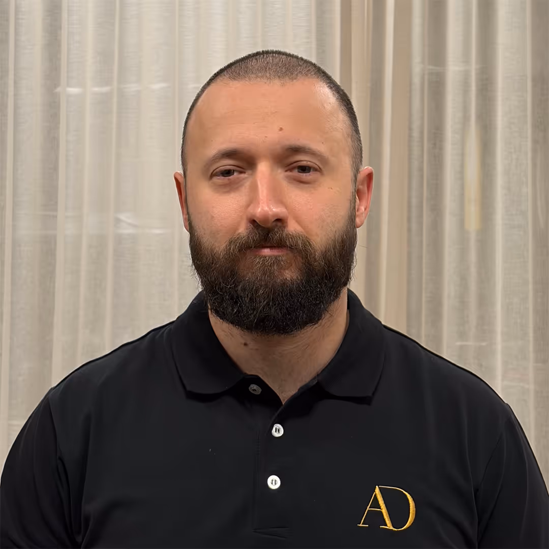 Glimlachende man van middelbare leeftijd met zwart haar, bros, baard en snor, gekleed in een zwart poloshirt met gele AD-initialen, staande voor vitrages.