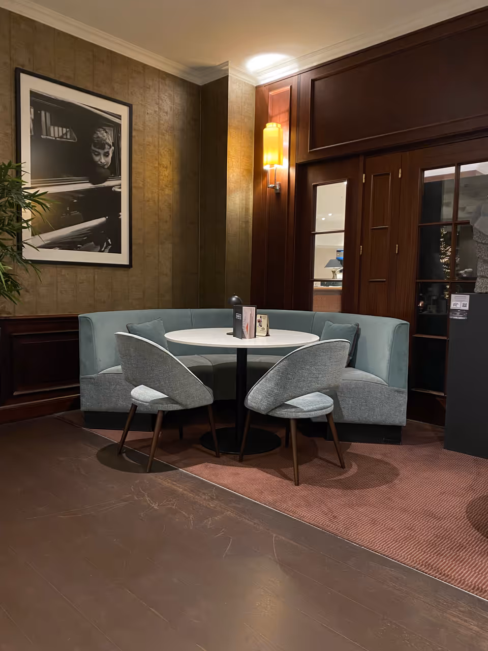 Gezellige zithoek met een ronde witte tafel, een gebogen blauwgroene bank, twee grijze stoelen en een zwart-witfoto aan de muur.