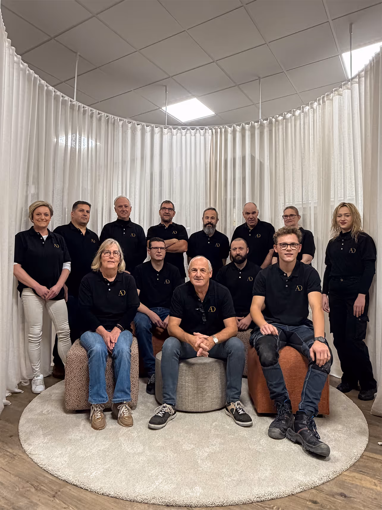 Een groep van dertien mensen in zwarte poloshirts met een gouden logo poseert binnenshuis op een beige tapijt met witte gordijnen op de achtergrond.