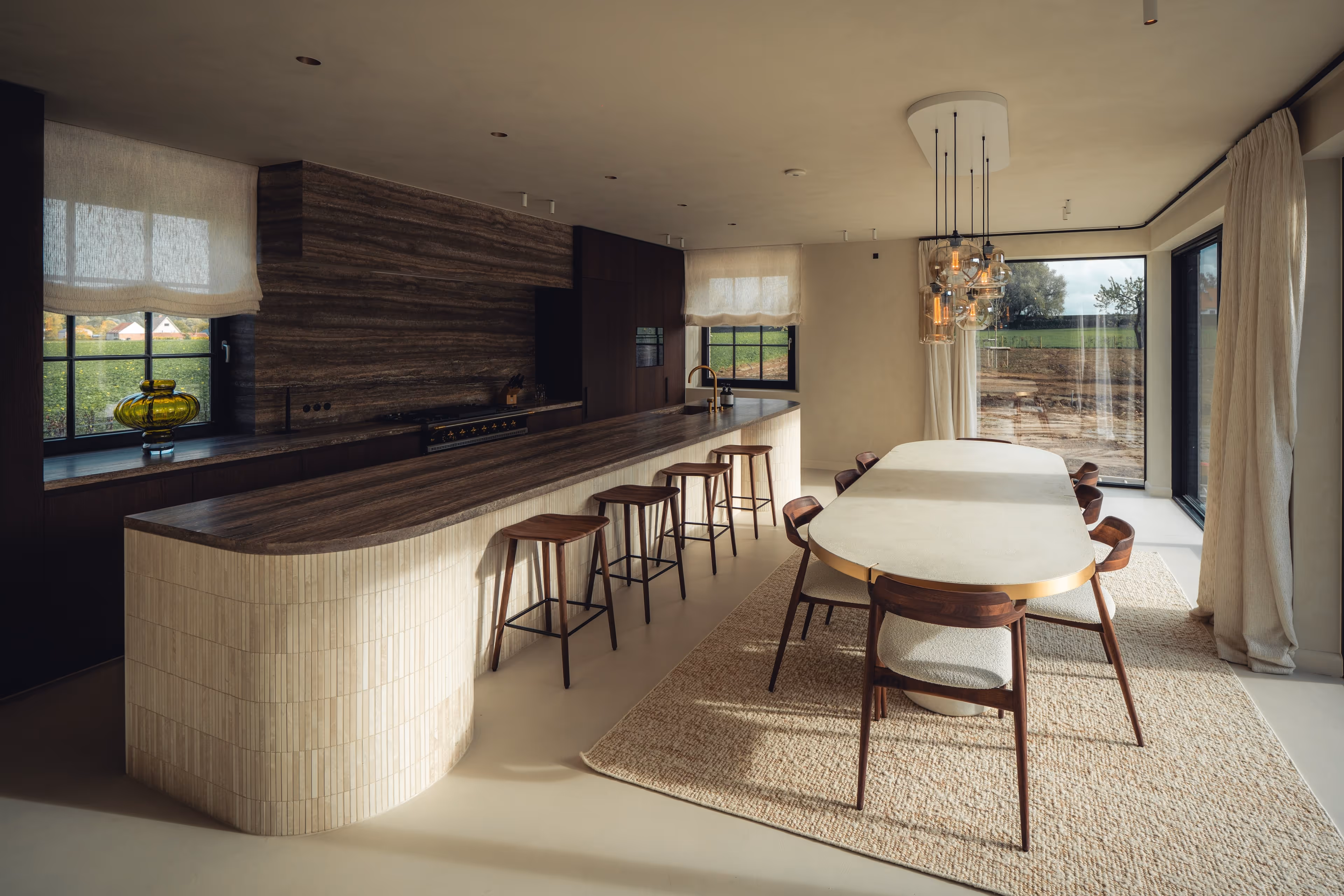 Moderne eetkamer met een grote ovale tafel, houten stoelen, een tapijt met structuur en een aangrenzend kookeiland met houten barkrukken en grote ramen.
