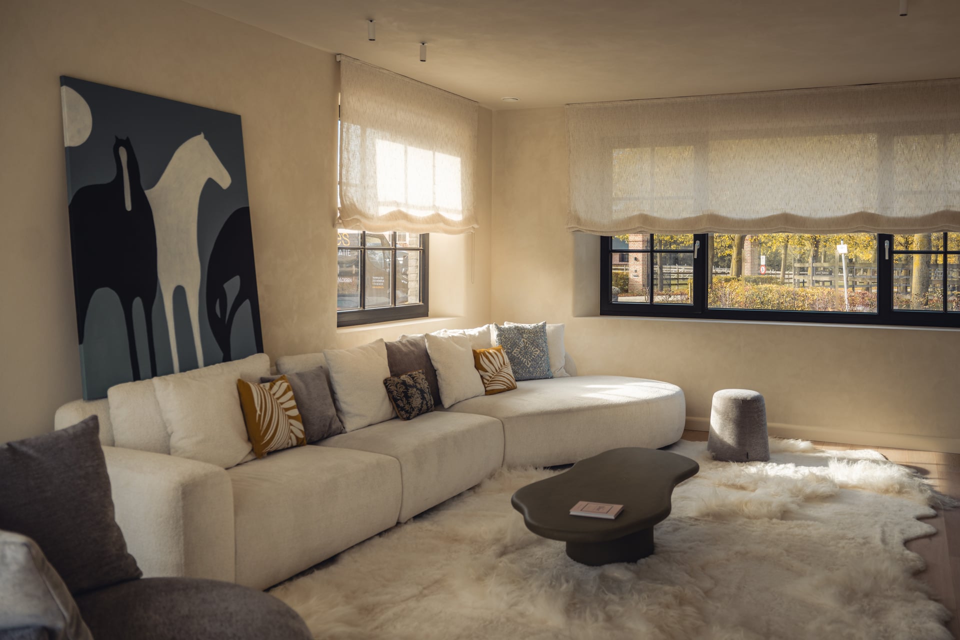 Lichtbeige, gestoffeerde zetel met organische rondingen; witte, okeren, bruine en blauwe kussens, een schilderij met paarden, zwarte ramen met vouwgordijnen, licht, donzig tapijt.