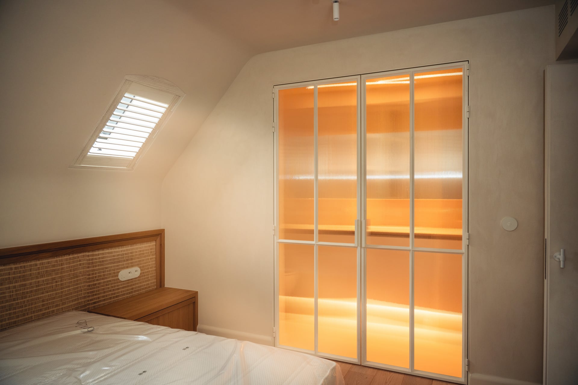 Slaapkamer met houten, gestoffeerd hoofdeinde, lichtbeige kamer, dakraam met witte jaloezieën, verlichte ruimte achter glazen deuren.