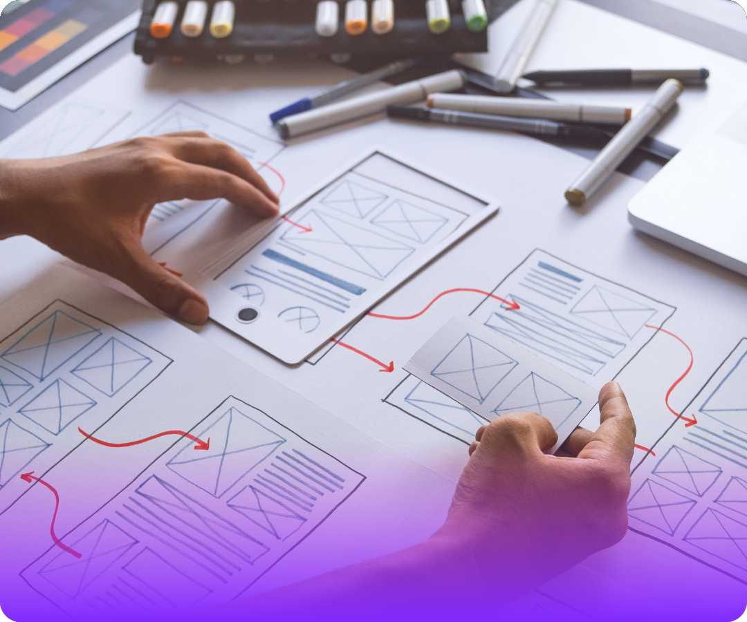 Hands arranging wireframe sketches connected by red arrows for website or app UI design on a desk with markers.