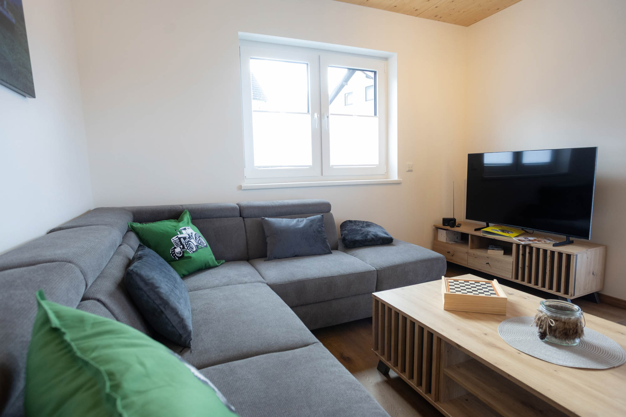 Wohnzimmer mit grauem Ecksofa, grünen und grauen Kissen, hölzernem Couchtisch mit Schachbrett und TV auf einem TV-Schrank.