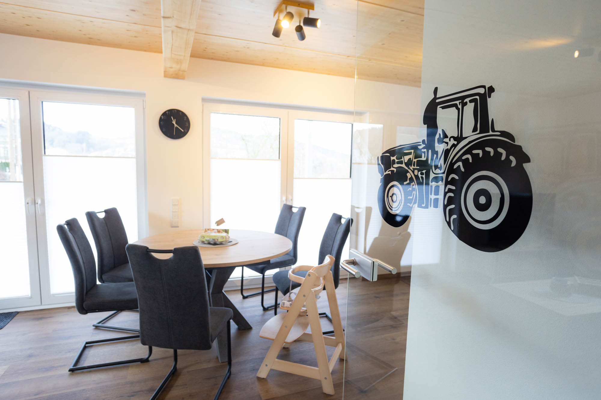 Esszimmer mit einem runden Holztisch, sechs grauen Stühlen und einem Kinderhochstuhl vor großen Fenstern, an der Glastür ein schwarzer Aufkleber mit einem Traktor.