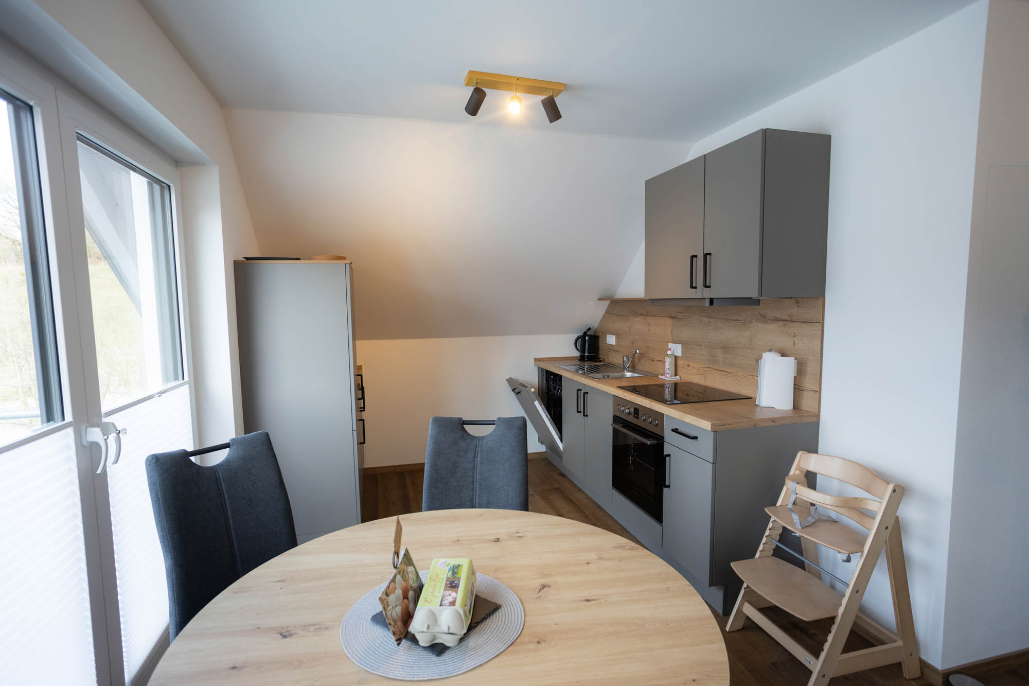 Moderne Küche mit grauen Schränken, runden Holztisch mit zwei dunkelgrauen Stühlen, einem Kinderhochstuhl und einer großen Fensterfront.