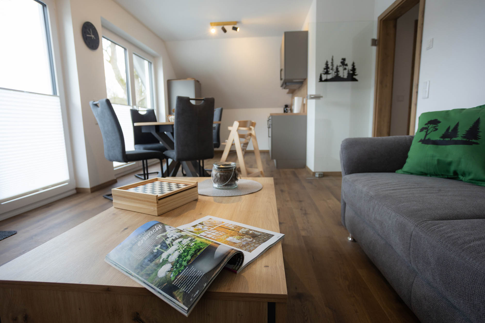 Moderner Wohnraum mit grauem Sofa, grünem Kissen mit Waldmotiv, hölzernem Couchtisch mit offenem Magazin und Schachspiel, Esstisch mit Stühlen und Küche im Hintergrund.